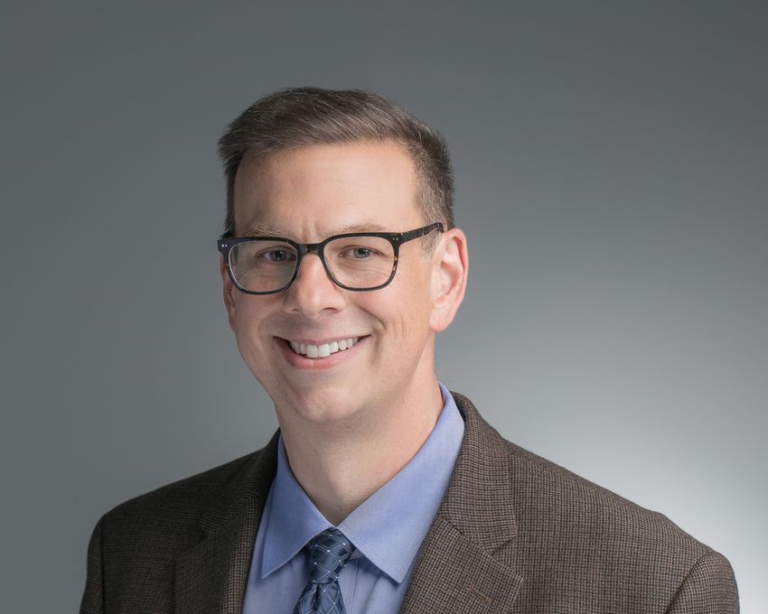 Matthew Girgenti, wearing glasses, a blue shirt, a patterned dark blue tie, and a dark brown patterned suit, poses in front of a grey background. Matthew Girgenti, wearing glasses, a blue shirt, a patterned dark blue tie, and a dark brown patterned suit, poses in front of a grey background.